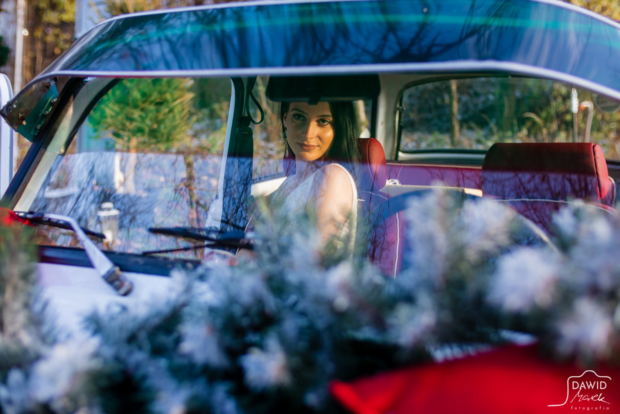 Panna młoda w białej sukni siedzi w czerwonym, klasycznym samochodzie. Dekoracje świąteczne na przednim planie. Unikalne ujęcie przez szybę z refleksami.