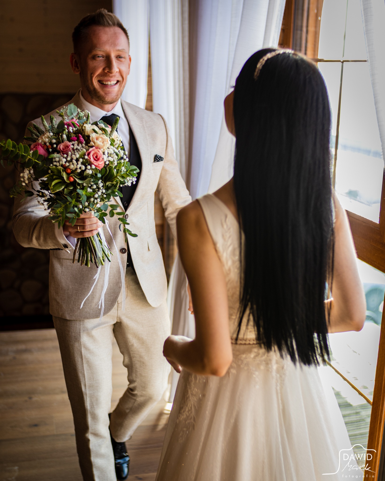 Uśmiechnięty pan młody w beżowym garniturze trzyma bukiet kwiatów, patrząc na pannę młodą w białej sukni stojącą przy oknie.