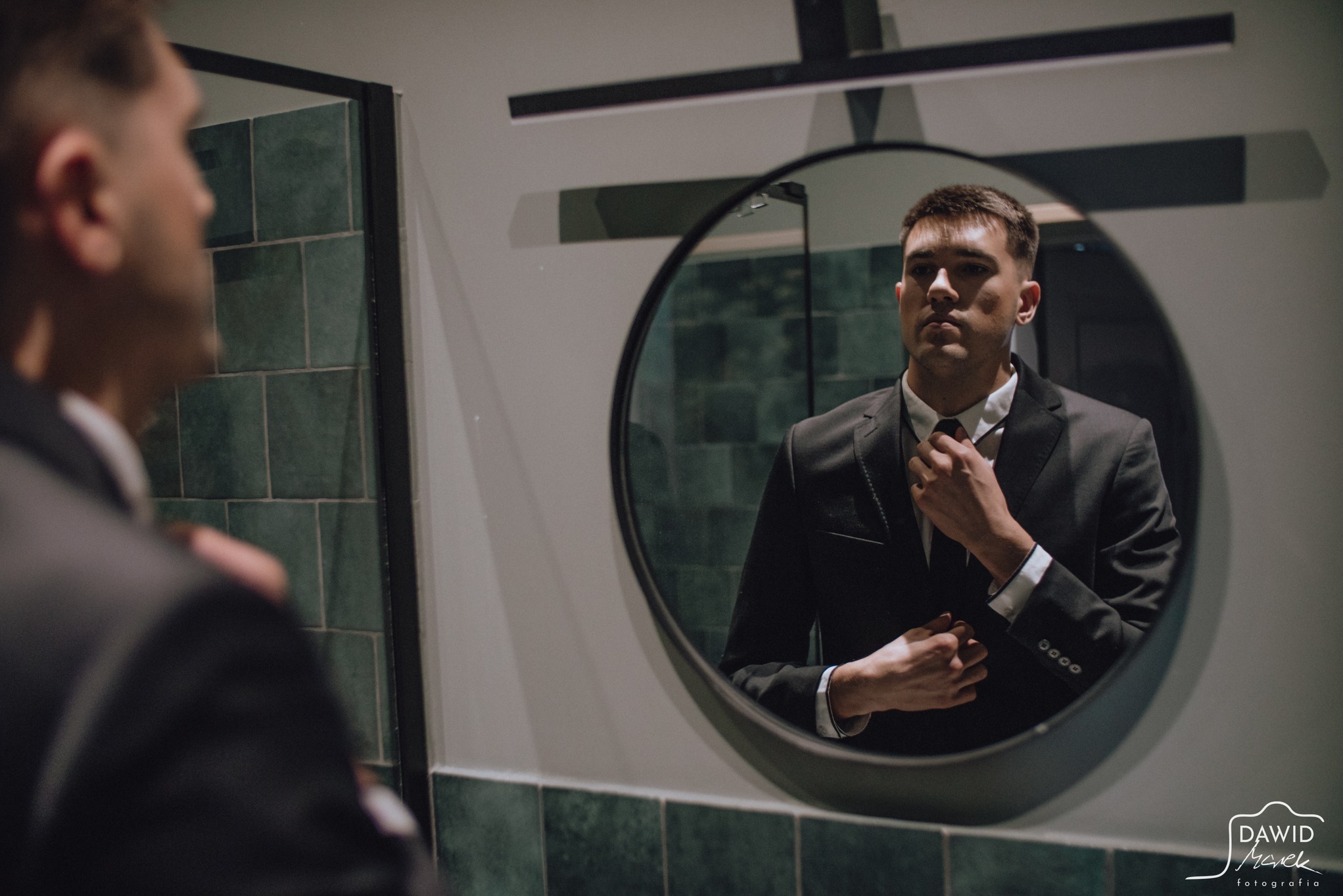 Mężczyzna w garniturze poprawia krawat, widoczny w okrągłym lustrze w łazience z zielonymi płytkami. Odbicie jego sylwetki po lewej stronie kadru.