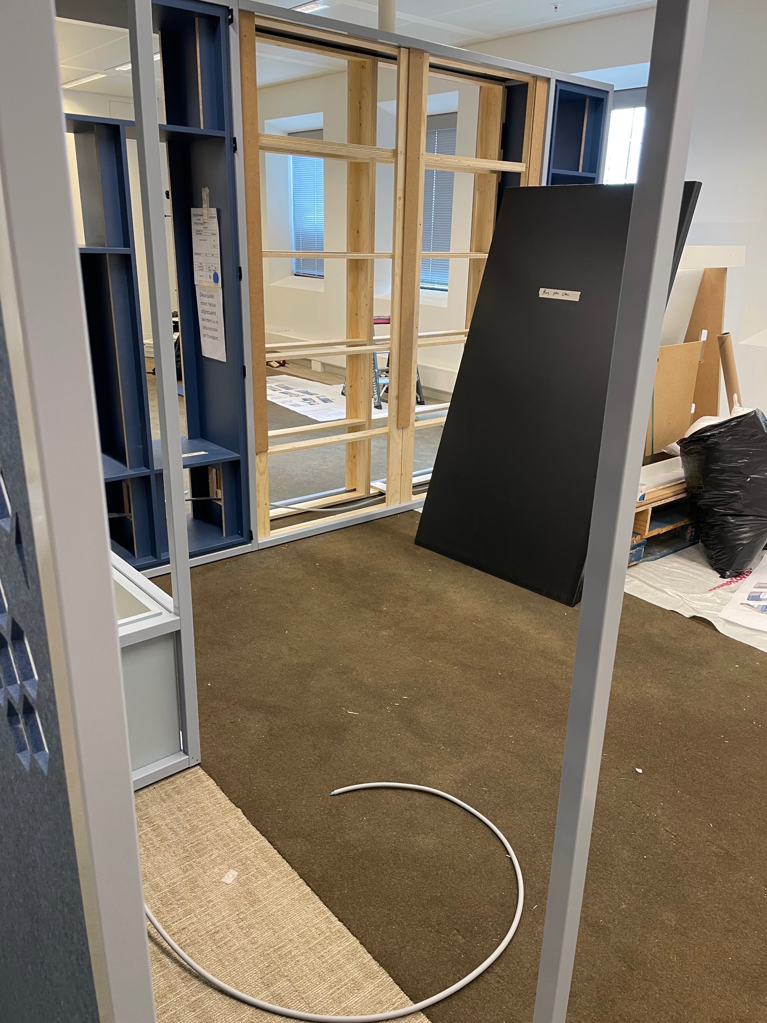 Wnętrze biura w trakcie montażu ścianek działowych z drewnianą konstrukcją i niebieskimi elementami, widoczne kable i śmieci, brązowa wykładzina dywanowa.