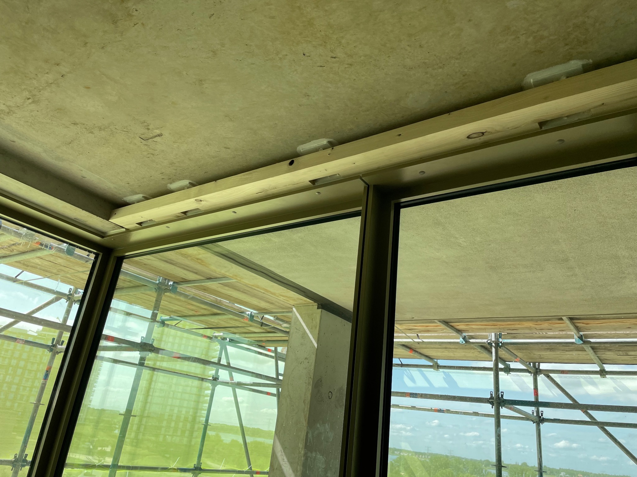 Montaż okna z widokiem na rusztowanie i krajobraz. Drewniane elementy konstrukcyjne przy suficie, surowy beton. Widok z wnętrza budynku w trakcie wykończenia.