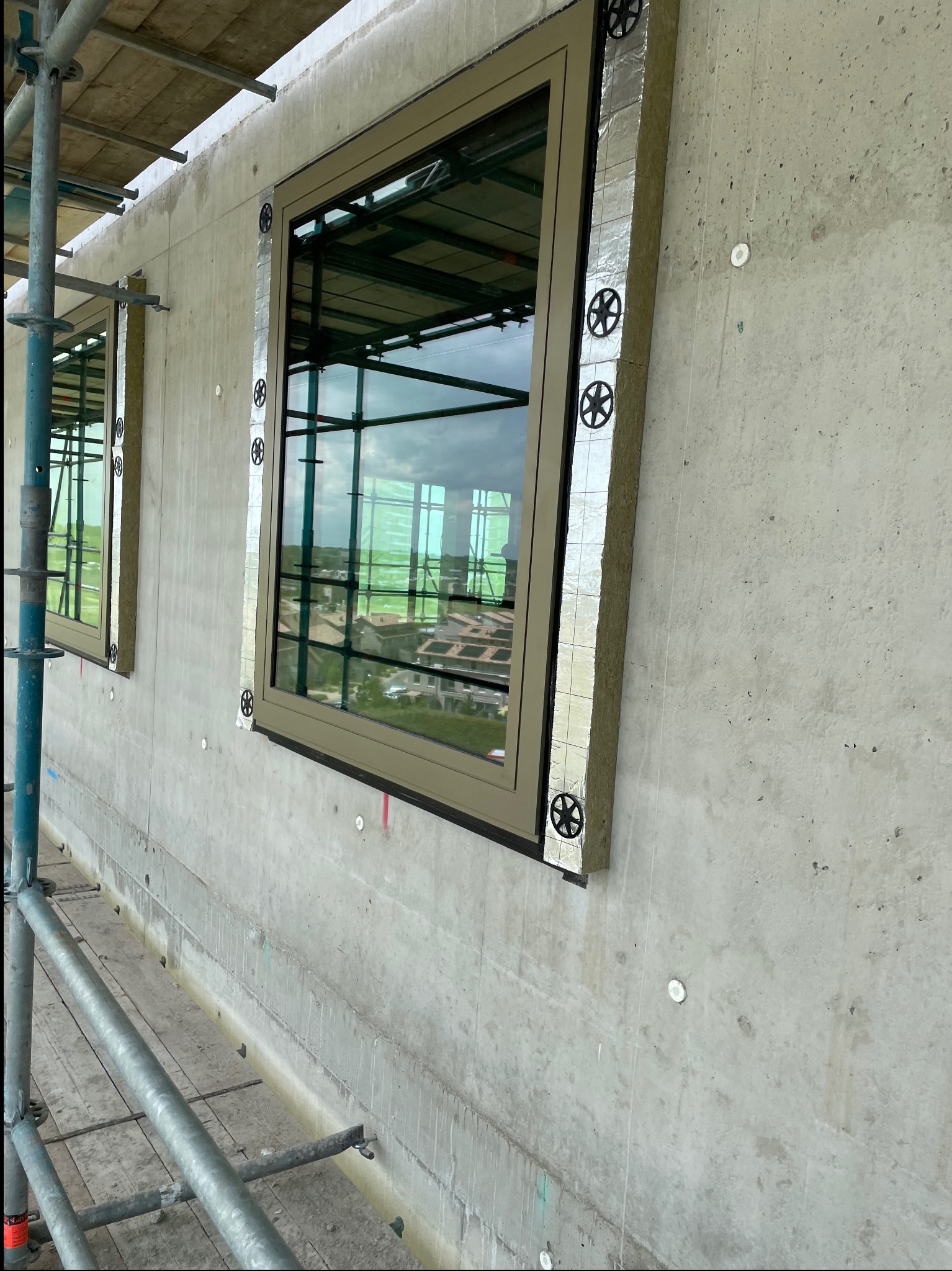 Montaż okien w surowej ścianie budynku. Widoczne ocieplenie wokół ram okiennych, rusztowanie i betonowa powierzchnia ściany z widocznymi elementami mocującymi.