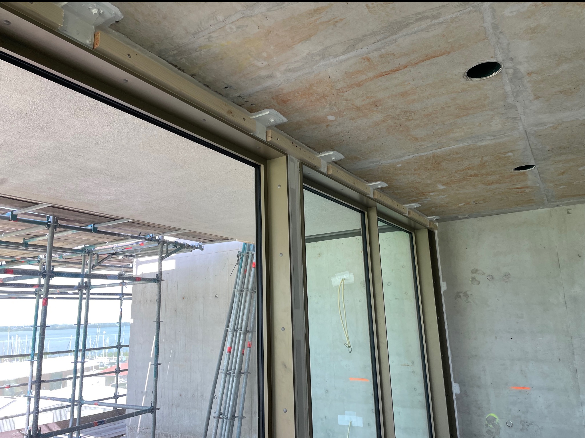 Surowy stan wnętrza z oknami, widoczne metalowe wsporniki mocujące do sufitu, rusztowania na zewnątrz okna. Prace wykończeniowe w toku.