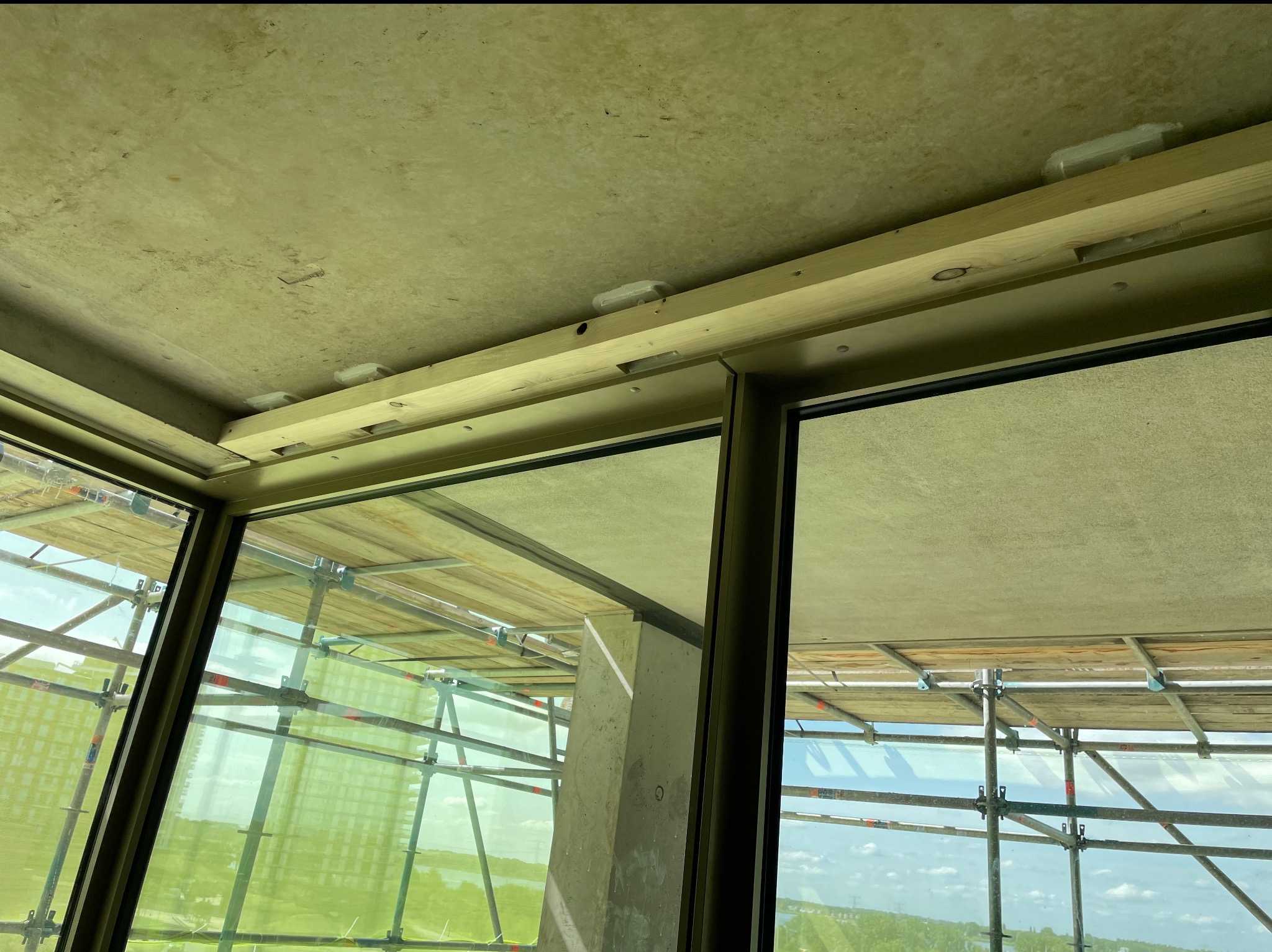 Surowy strop z drewnianą ramą okienną i widokiem na rusztowania. W tle budynki i krajobraz. Widać elementy montażowe.