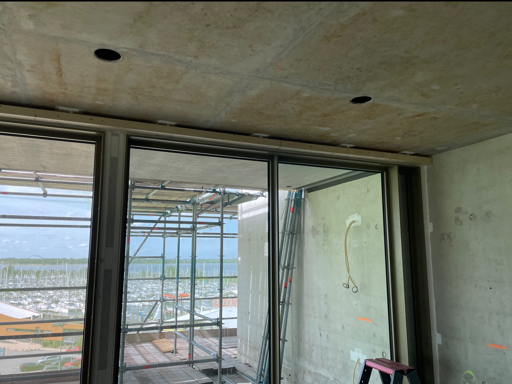 Surowy sufit z widocznymi otworami pod oświetlenie i drewnianą listwą, duże okna z widokiem na przystań jachtową, rusztowania na zewnątrz. Wnętrze w trakcie wykończenia.