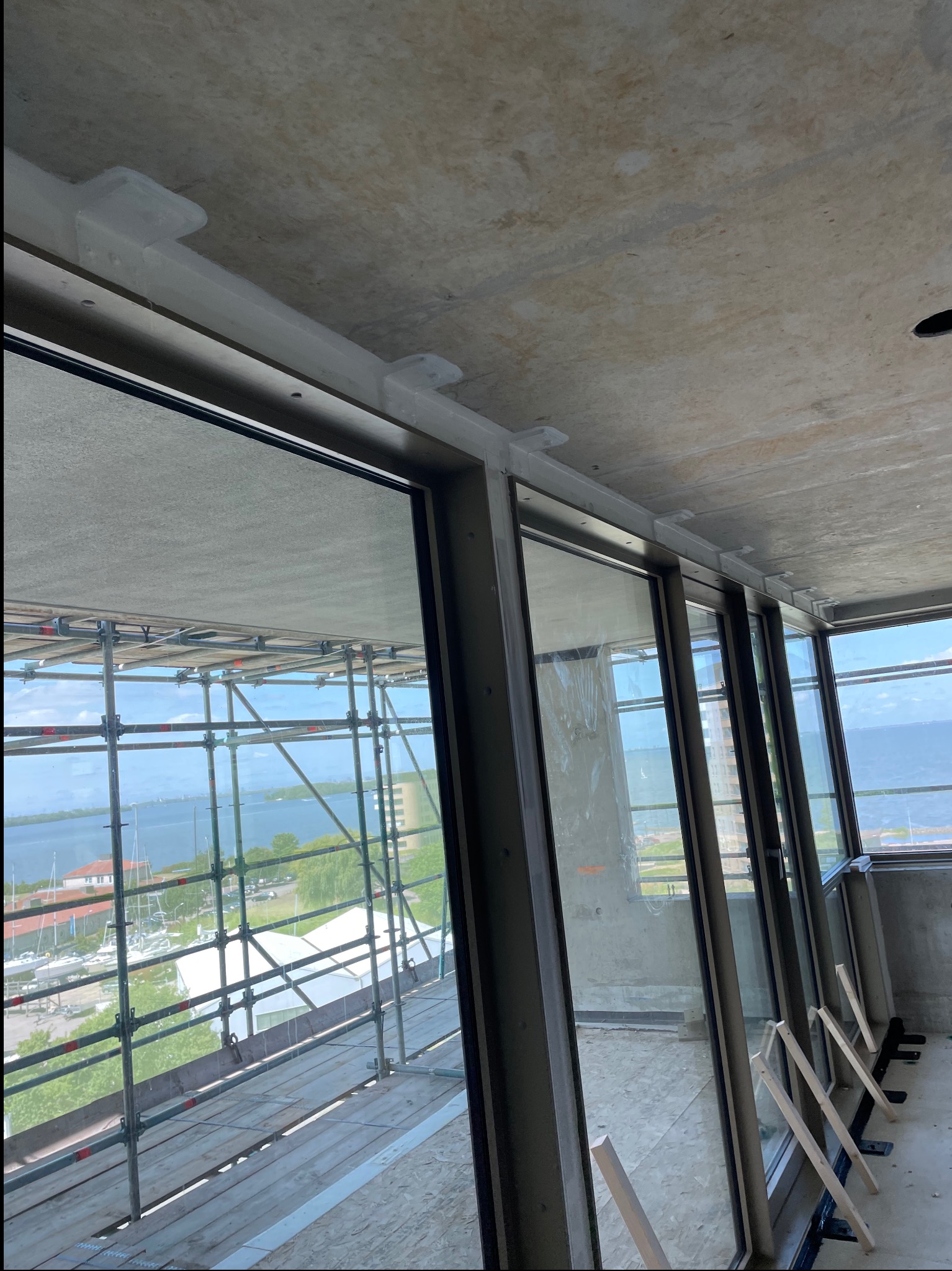 Montaż okien przesuwnych w surowym stanie. Widoczne szyny mocujące, surowy betonowy strop i rusztowanie za oknami z widokiem na jezioro.