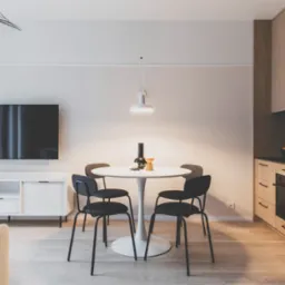 Jasne wnętrze: okrągły stół z czarnymi krzesłami, minimalistyczna szafka RTV z telewizorem i drewniana zabudowa kuchenna z czarnym blatem. Nowoczesny design.