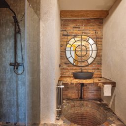 Lucyna Joskowska - Industrialna łazienka: ceglana ściana z okrągłym lustrem, drewniany blat z czarną umywalką, kabina prysznicowa z ciemną armaturą i nietypowa, ceglana wanna.