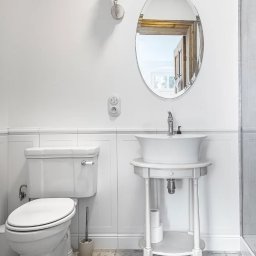 Lucyna Joskowska - Jasna łazienka w stylu retro z owalnym lustrem, toaletą i umywalką na ozdobnym stoliku. Uwagę zwraca lampa ścienna z abażurem i wzorzysta podłoga.