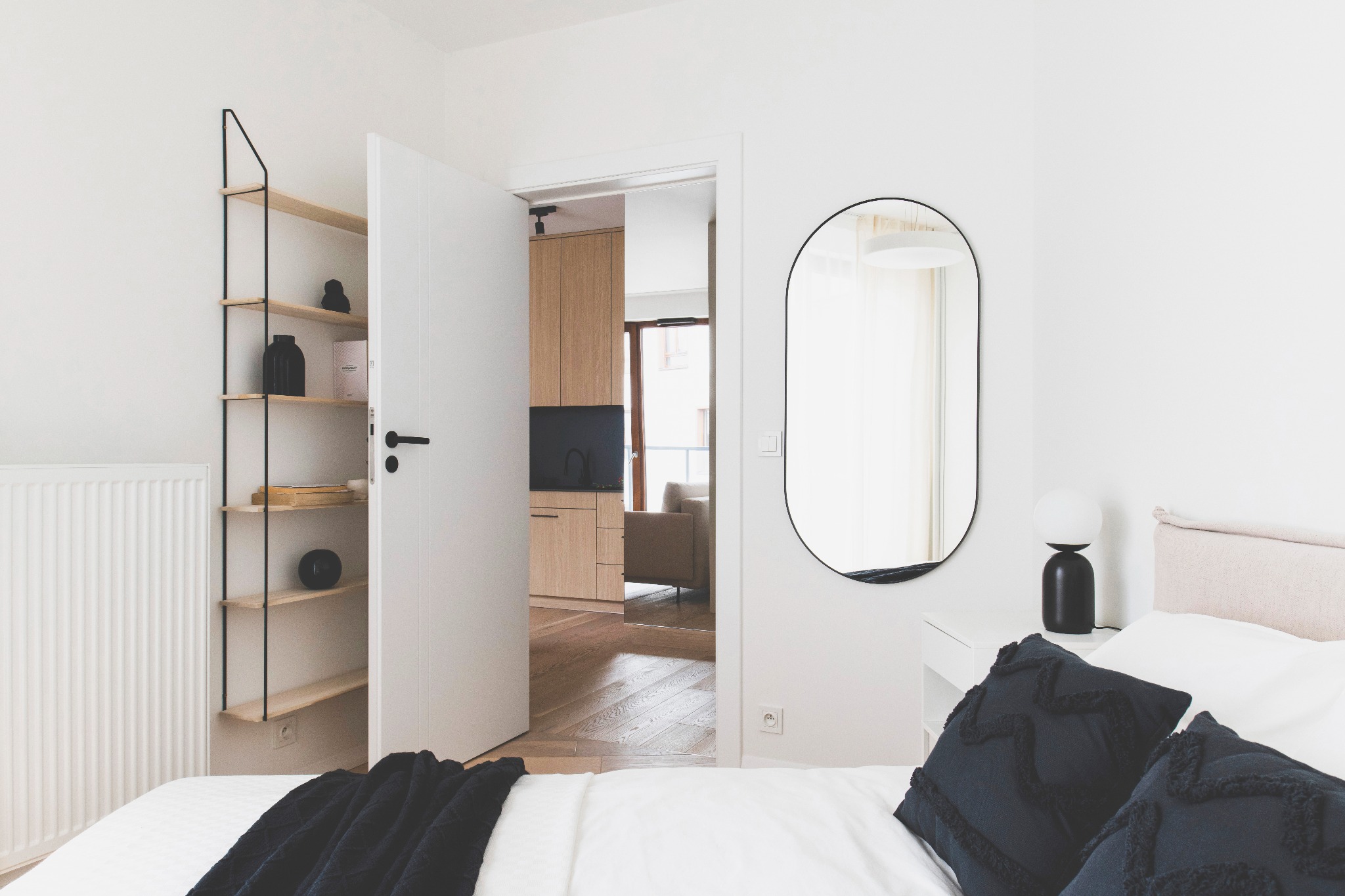 Jasna sypialnia z minimalistycznym regałem, owalnym lustrem i otwartymi drzwiami do salonu z drewnianą podłogą i meblami. Nowoczesny design wnętrza.