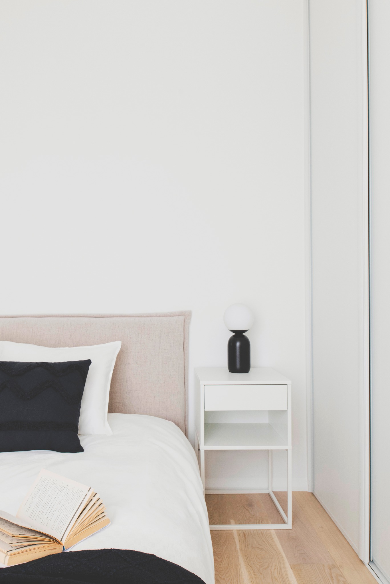 Minimalistyczna sypialnia: beżowe wezgłowie łóżka, czarna poduszka z wzorem, biała pościel, stolik nocny z lampką i otwarta książka na łóżku.