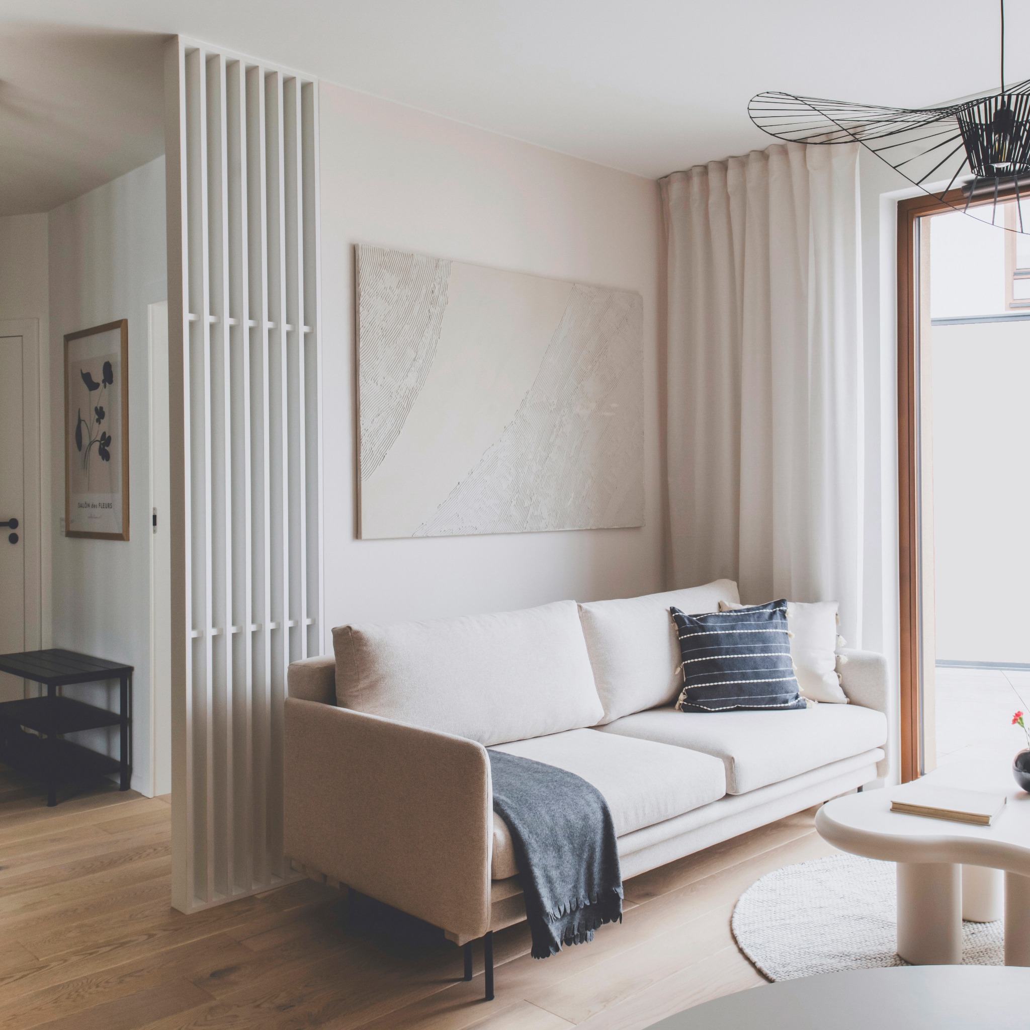 Jasny salon z beżową sofą, abstrakcyjnym obrazem i drewnianą podłogą. Biały parawan lamelowy oddziela przestrzeń. Widoczny stolik kawowy i okno z zasłonami.