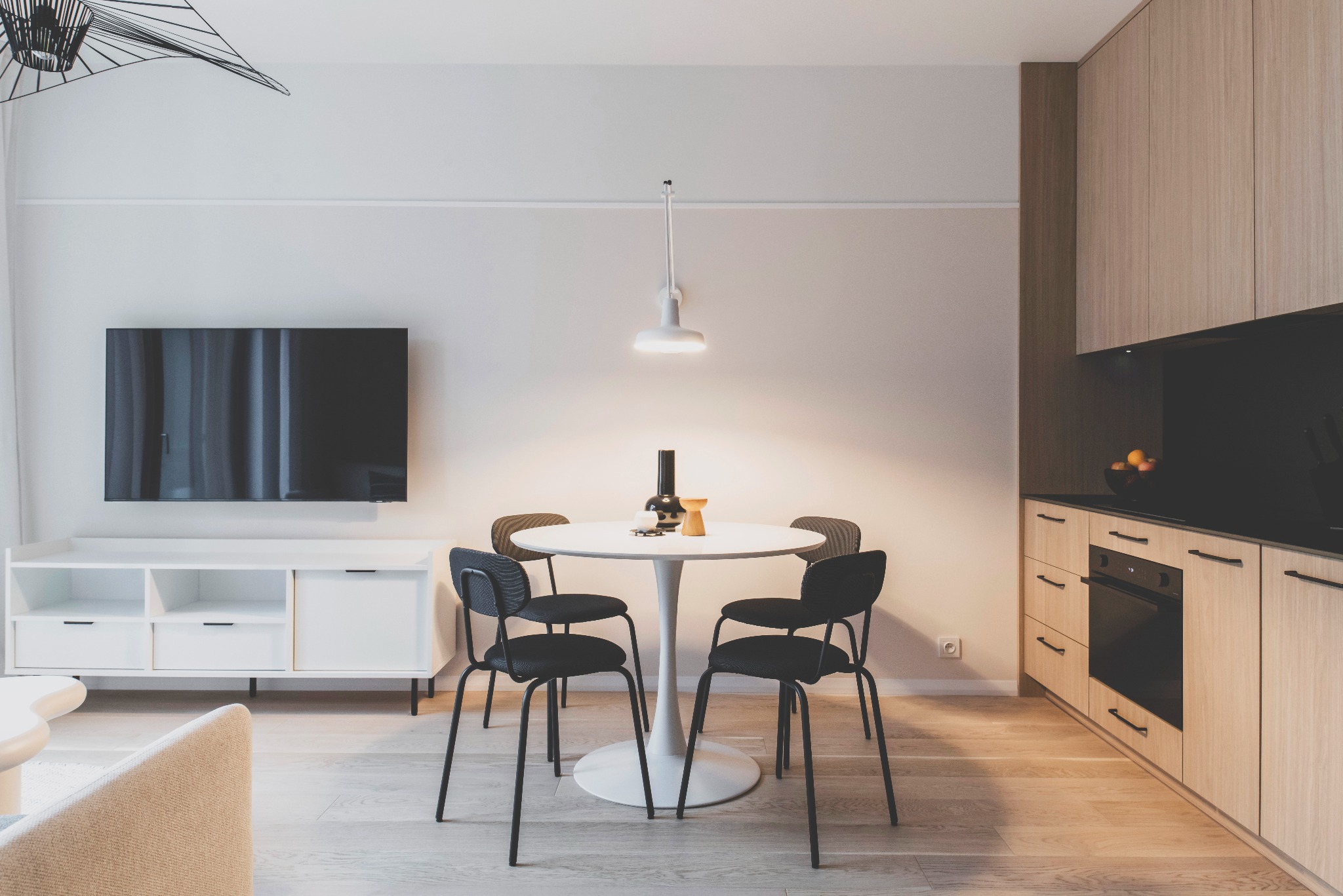 Jasne wnętrze: okrągły stół z czarnymi krzesłami, minimalistyczna szafka RTV z telewizorem i drewniana zabudowa kuchenna z czarnym blatem. Nowoczesny design.