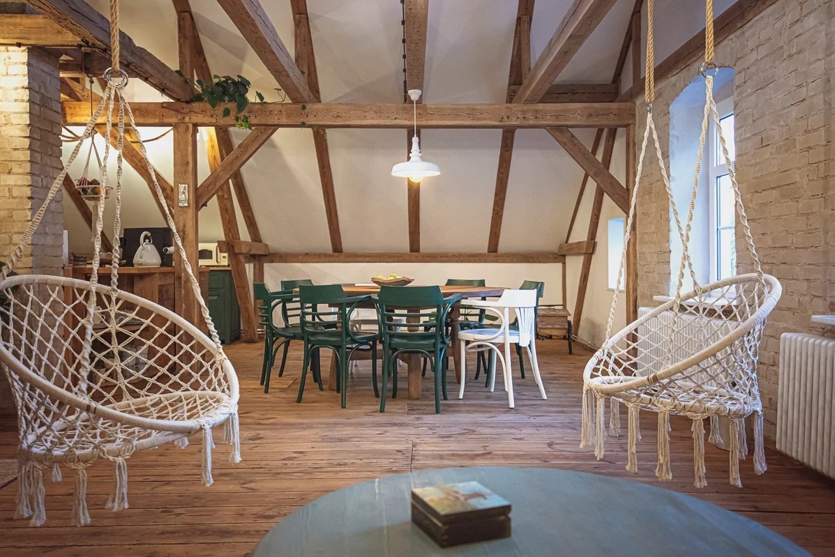 Rustykalne wnętrze z drewnianą konstrukcją stropu, wiszącymi fotelami makrama i stołem z krzesłami. Ceglana ściana i drewniana podłoga dodają ciepła.