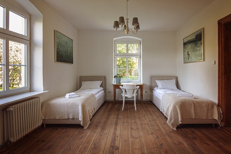 Jasna sypialnia z dwoma łóżkami pojedynczymi, drewnianą podłogą i oknem z widokiem na zieleń. Na ścianach obrazy w ramach i żyrandol.