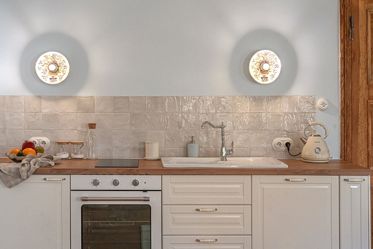 Jasna kuchnia w stylu rustykalnym: kremowe szafki, drewniany blat, ceramiczne płytki imitujące starą cegłę, dekoracyjne lampy ścienne i owoce w misie.