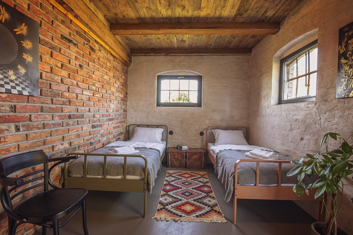 Rustykalna sypialnia z ceglaną ścianą, drewnianym sufitem i dwoma łóżkami. Na ścianie wiszą obrazy, a na podłodze leży wzorzysty dywan.