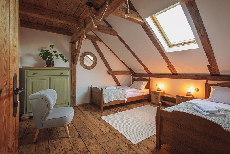 Pokój na poddaszu z drewnianymi belkami, dwoma łóżkami, szafkami nocnymi z lampkami, zieloną komodą i fotelem. Okrągłe lustro i dywan dodają przytulności.