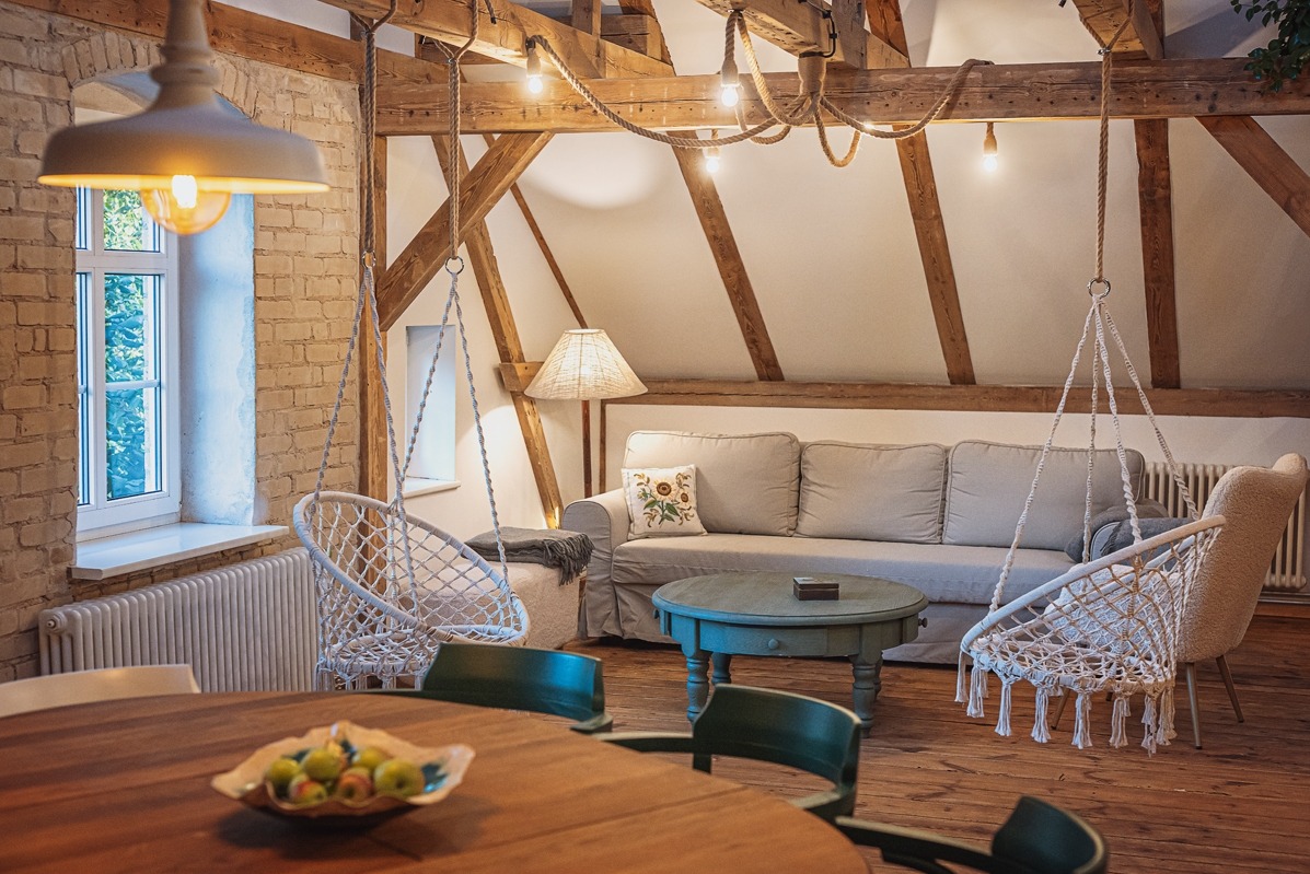 Stylowa aranżacja poddasza w Gdańsku: drewniane belki, wiszące fotele, okrągły stół z owocami, beżowa sofa i lampa. Przytulne i unikalne wnętrze.