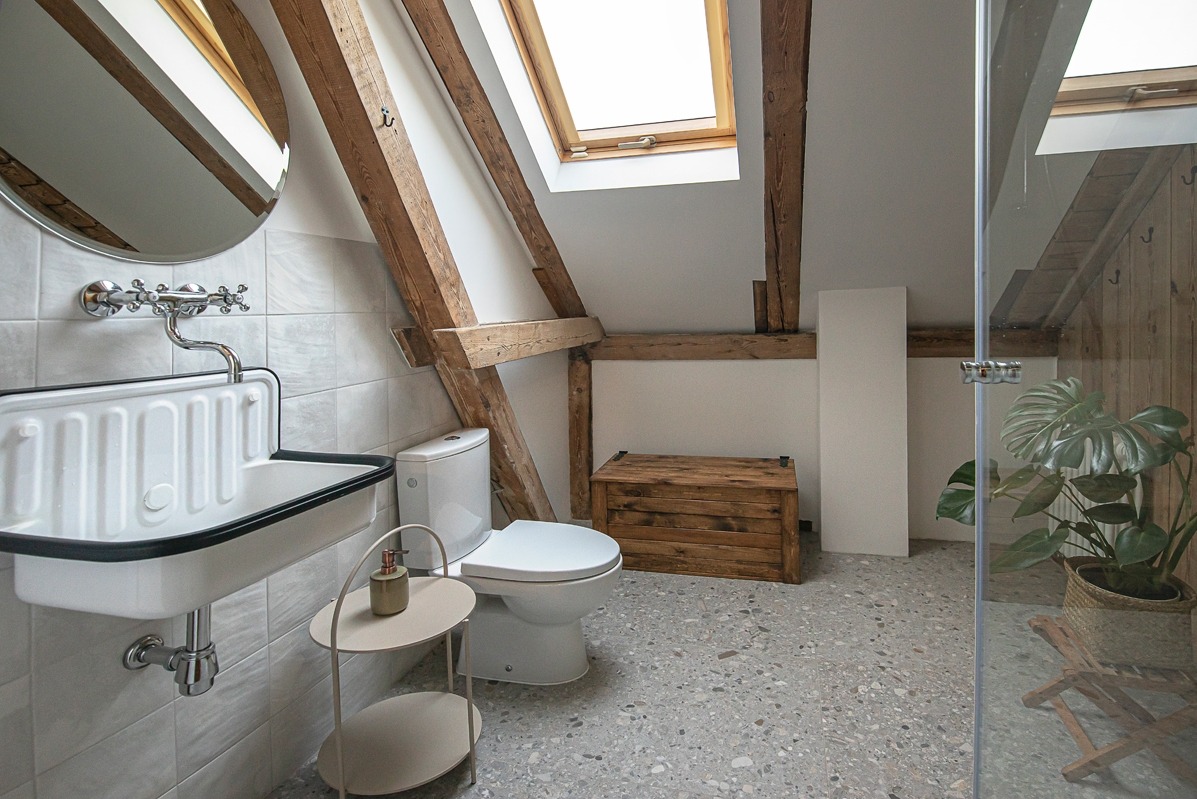 Stylowa łazienka na poddaszu z drewnianymi belkami, białą ceramiką i szarą mozaiką. Uwagę przykuwa retro umywalka i skrzynia z surowego drewna.