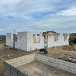 Wznoszenie ścian domu z bloczków betonu komórkowego. Widoczny murarz na rusztowaniu, taczka, agregat prądotwórczy i przyczepa. W tle krajobraz i błękitne niebo.