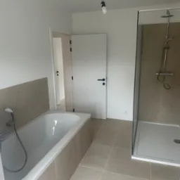 Nowoczesna łazienka po remoncie: wanna z baterią, przeszklona kabina prysznicowa, beżowe płytki podłogowe i ścienne, białe ściany i drzwi.