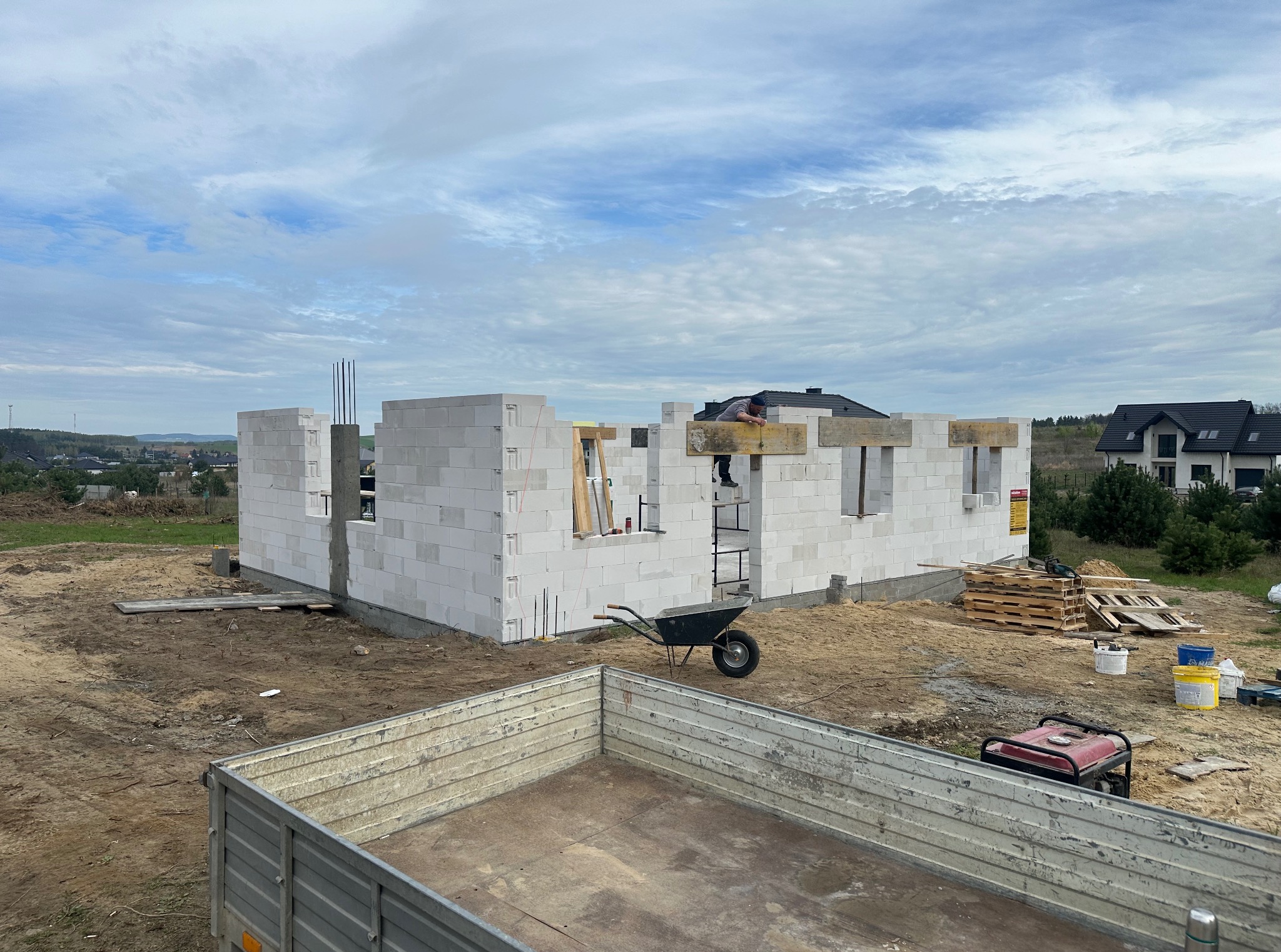 Wznoszenie ścian domu z bloczków betonu komórkowego. Widoczny murarz na rusztowaniu, taczka, agregat prądotwórczy i przyczepa. W tle krajobraz i błękitne niebo.