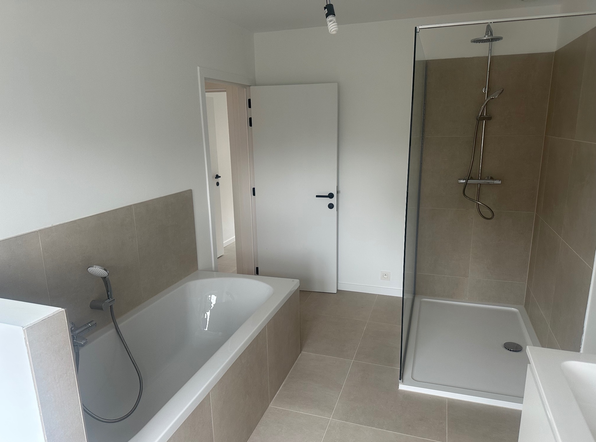 Nowoczesna łazienka po remoncie: wanna z baterią, przeszklona kabina prysznicowa, beżowe płytki podłogowe i ścienne, białe ściany i drzwi.