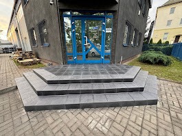 Nowoczesne schody wejściowe z szarych płytek prowadzące do biura z niebieskimi drzwiami. Widoczne elementy otoczenia i palety z boku.