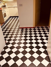 Ukończona podłoga w kuchni, wzór szachownicy z czarno-białych płytek ceramicznych. Widok z perspektywy wejścia, w tle jasne szafki kuchenne.