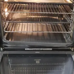 Wnętrze piekarnika Electrolux z wysuwanymi półkami i tackami, widoczne ustawienia temperatury na panelu sterowania. Piekarnik otwarty, gotowy do użycia.