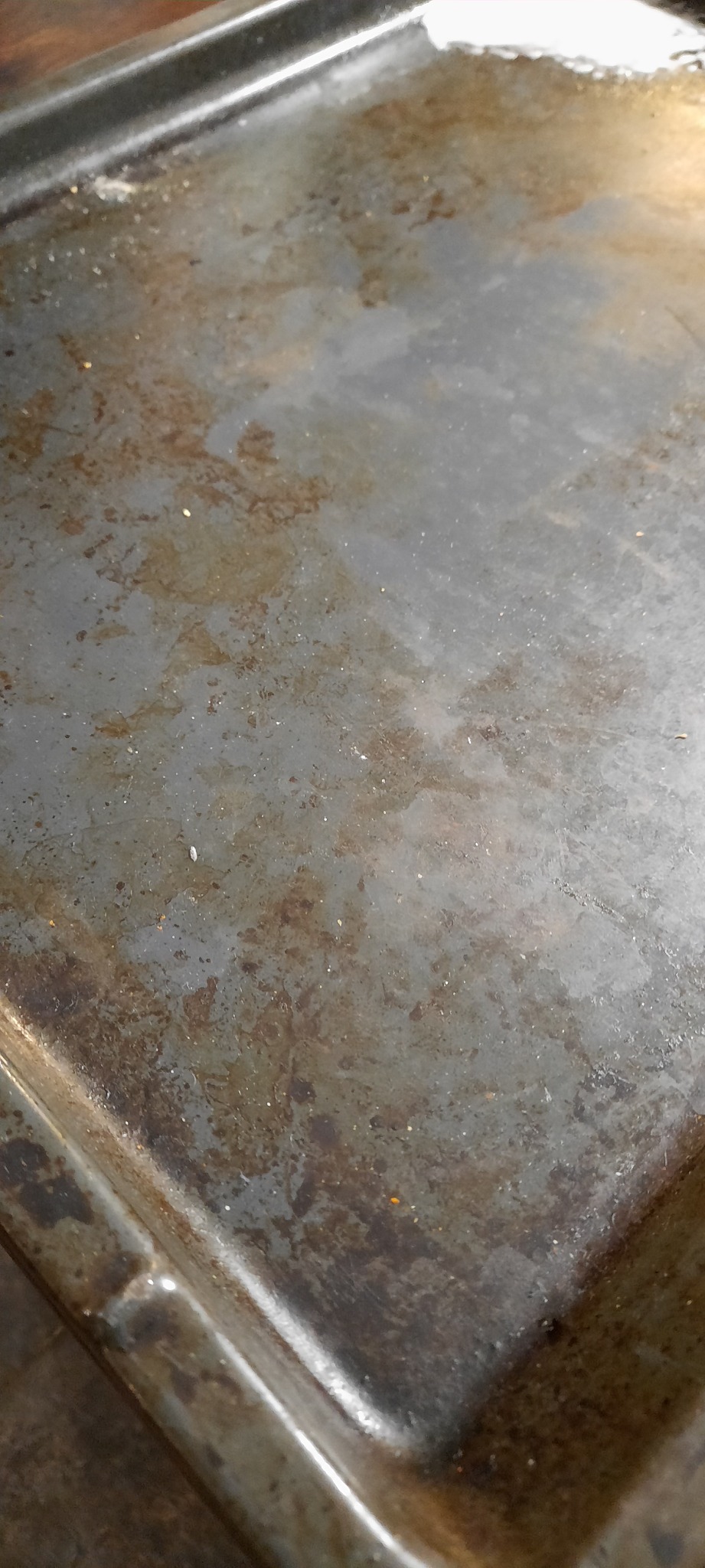 Brudna, metalowa blacha do pieczenia z przypalonymi resztkami i plamami. Widoczne drobne okruchy i zarysowania na powierzchni. Oświetlenie naturalne.