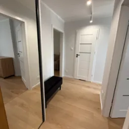 Wnętrze korytarza po sprzątaniu: jasne ściany, drewnopodobna podłoga, czarna ławka, białe drzwi z czarnymi akcentami, duże lustro z czarną ramą.