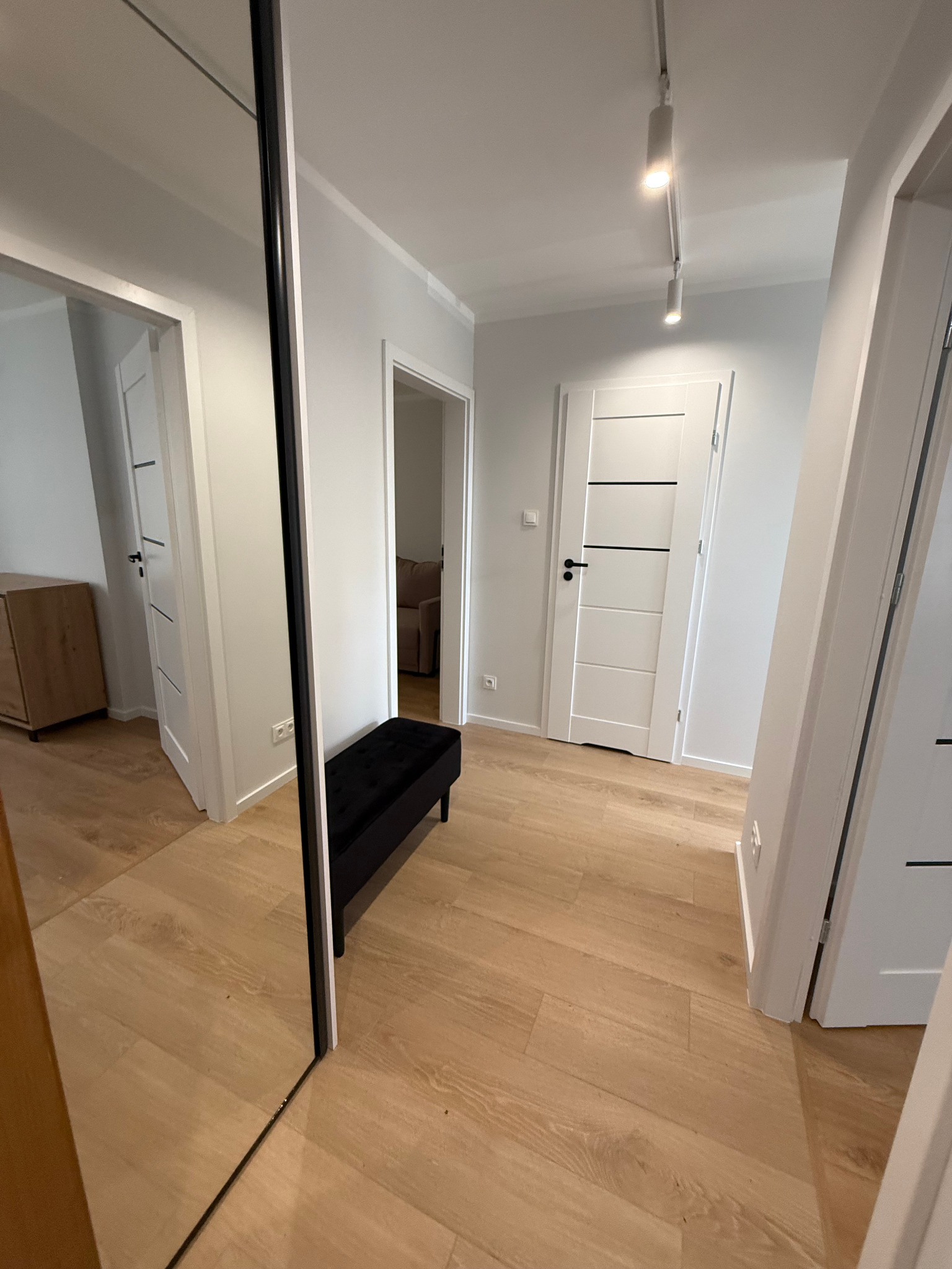 Wnętrze korytarza po sprzątaniu: jasne ściany, drewnopodobna podłoga, czarna ławka, białe drzwi z czarnymi akcentami, duże lustro z czarną ramą.