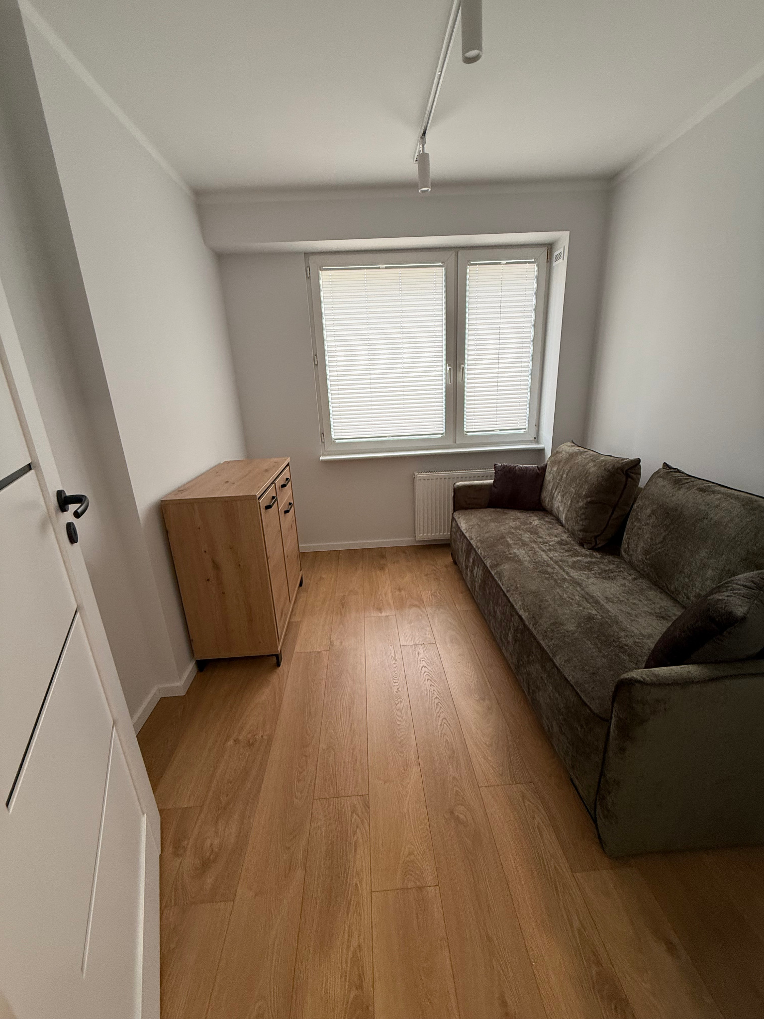 Jasne wnętrze pokoju po remoncie: drewniana podłoga, kanapa z poduszkami, komoda i okno z żaluzjami. Widok na czyste, odnowione pomieszczenie.