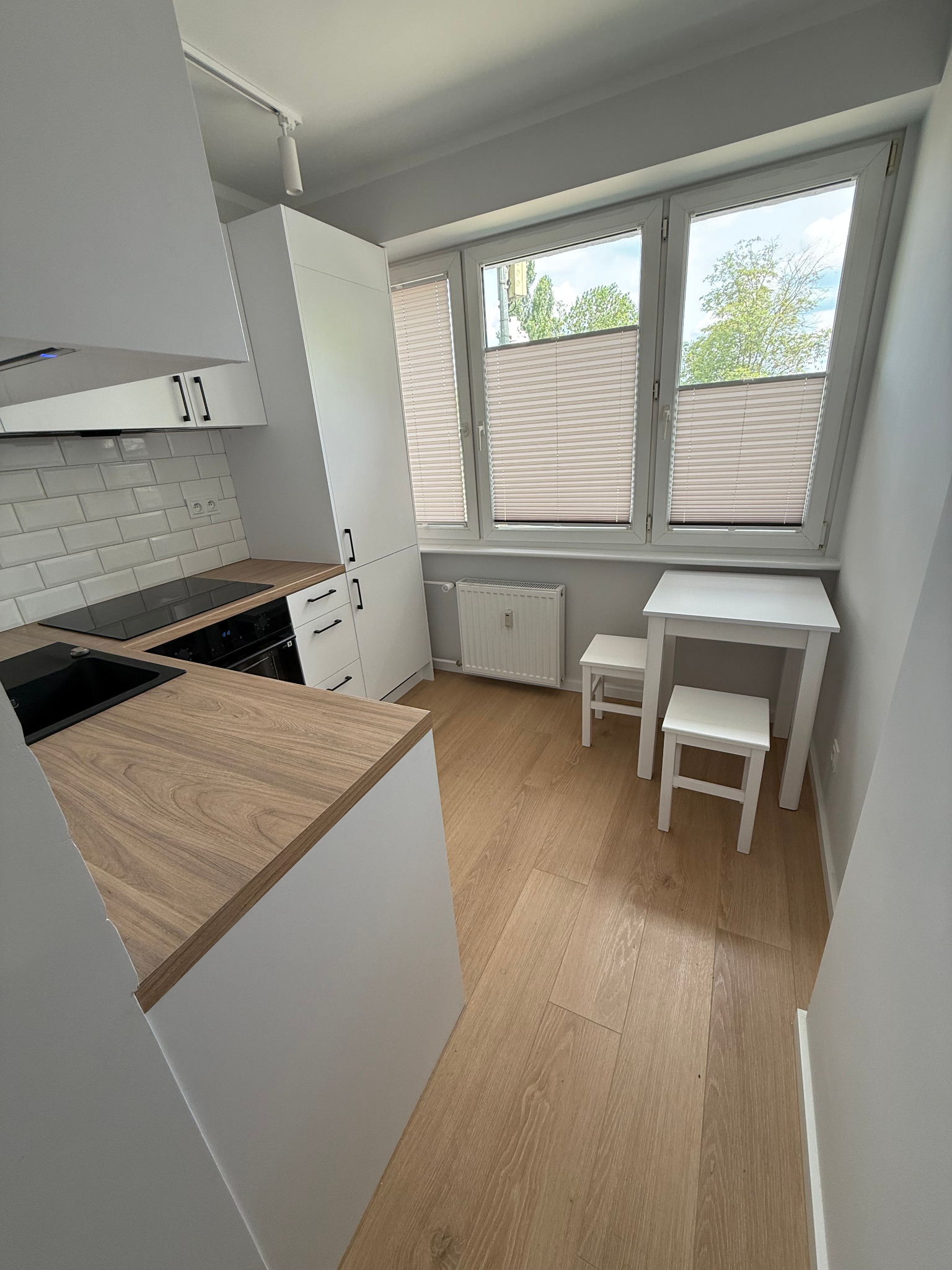 Jasna, nowoczesna kuchnia z drewnianym blatem i białymi meblami. Widoczne okno z żaluzjami, stół i krzesła. Minimalistyczny design, czyste linie, oświetlenie punktowe.