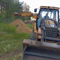 Damian (DS) - Żółta koparko-ładowarka CAT w trakcie pracy, nabiera ziemię z pryzmy. Widoczne trawy i drzewa w tle. W kabinie operator i dziecko.