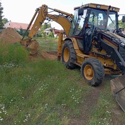 Damian (DS) - Żółta koparko-ładowarka CAT 428D w trakcie prac ziemnych, z widoczną stertą ziemi i wykopem na tle zieleni i zabudowań. Operator w kabinie.