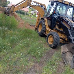 Damian (DS) - Żółta koparko-ładowarka CAT 428D podczas prac ziemnych, z widocznym wykopem w trawie z polnymi kwiatami. W tle domy mieszkalne i drzewa.