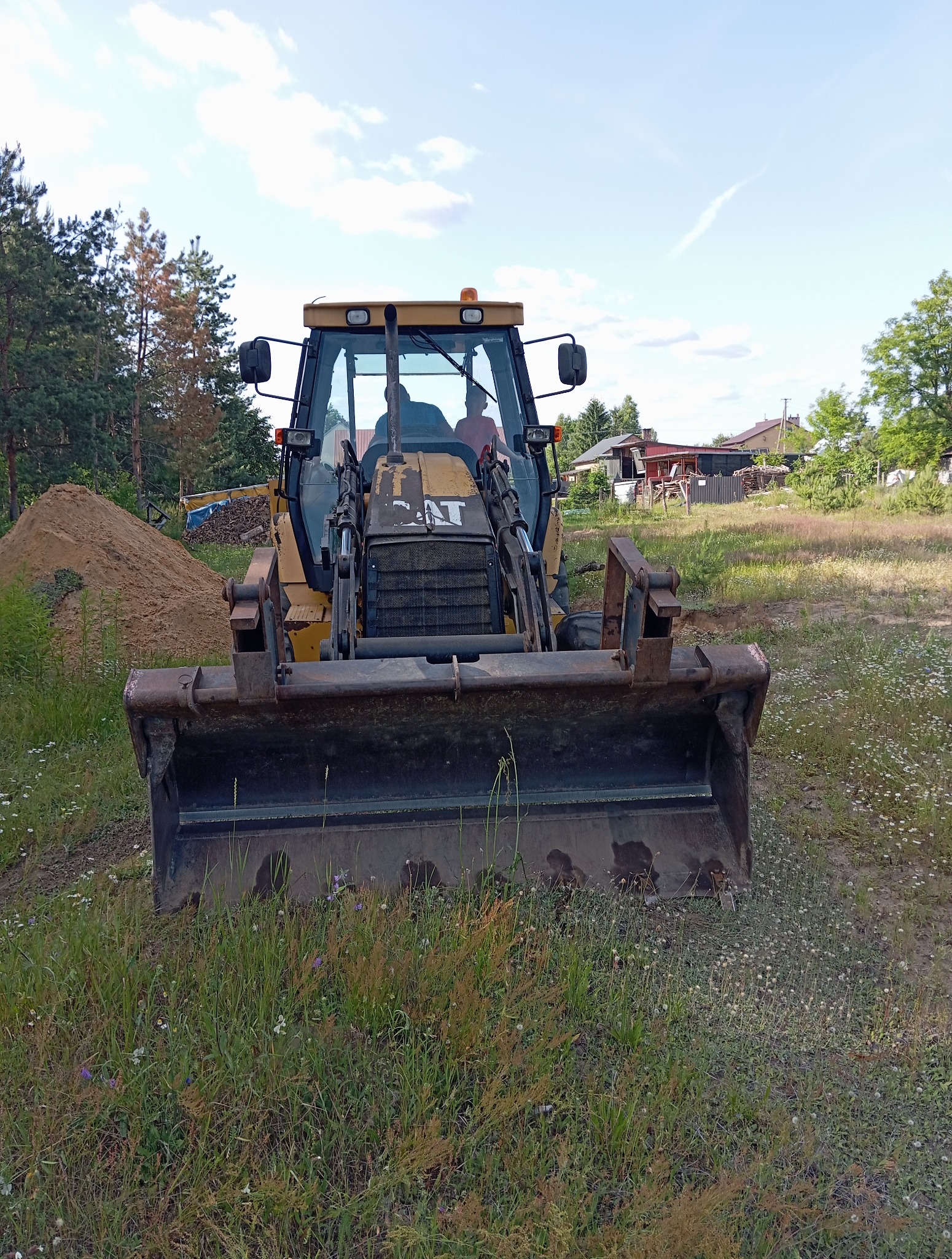 Żółta koparko-ładowarka CAT stoi na trawiastym terenie, z widocznym stosem ziemi w tle i zabudowaniami w oddali, sugerując prace ziemne.