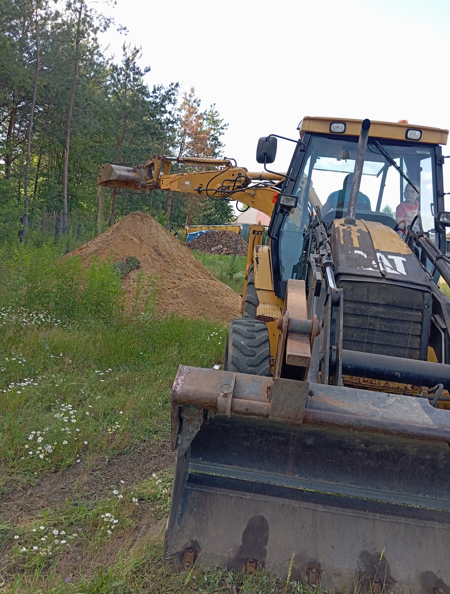 Żółta koparko-ładowarka CAT w trakcie pracy, nabiera ziemię z pryzmy. Widoczne trawy i drzewa w tle. W kabinie operator i dziecko.