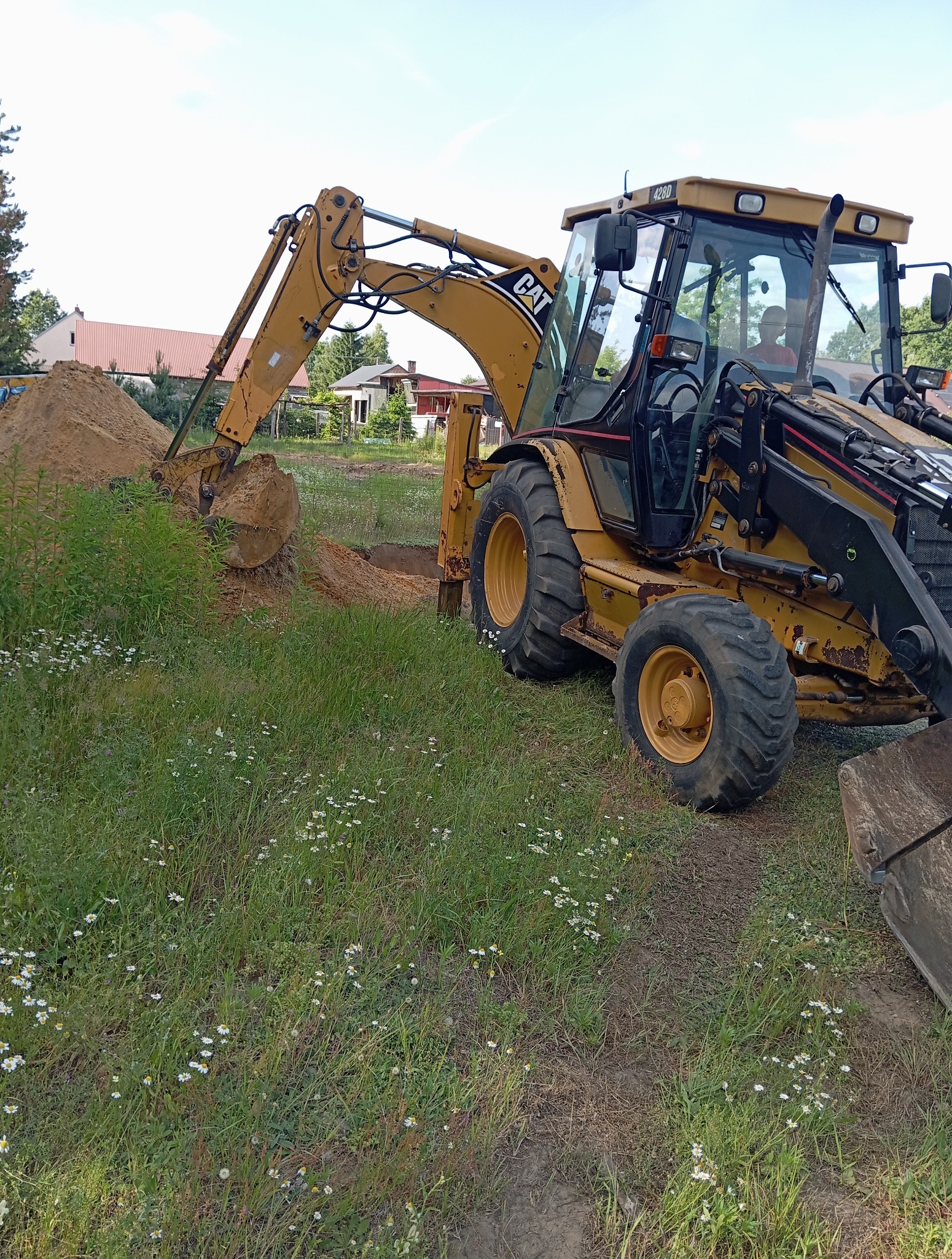 Żółta koparko-ładowarka CAT 428D w trakcie prac ziemnych, z widoczną stertą ziemi i wykopem na tle zieleni i zabudowań. Operator w kabinie.