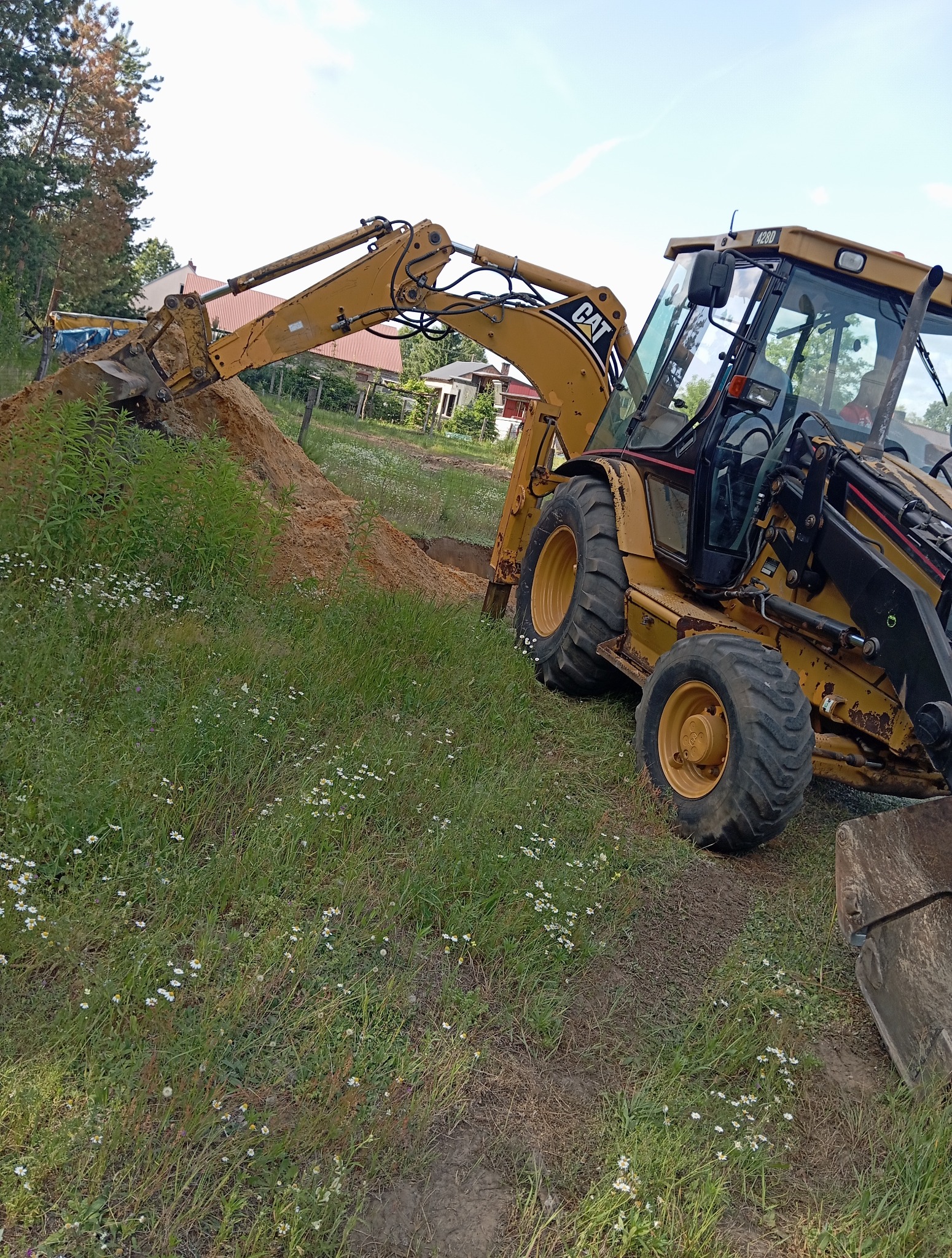 Żółta koparko-ładowarka CAT podczas wykopu w Pustkowie. Widok na maszynę w trawie z wykopaną ziemią w tle. Sprzęt budowlany w trakcie pracy.
