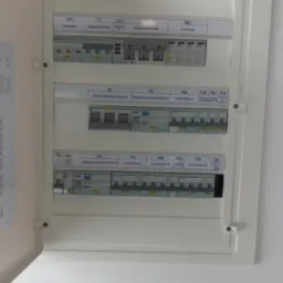 Otwarta skrzynka bezpiecznikowa z opisami i przełącznikami, widoczna na białej ścianie. Widok z bliska na elementy instalacji elektrycznej wewnątrz.