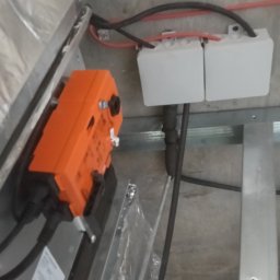 VoltMax - Pomarańczowy siłownik z przewodami i puszkami elektrycznymi zamontowany na metalowej konstrukcji pod sufitem. Widoczne kable i elementy instalacji.