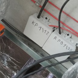 VoltMax - Dwa białe, podpisane puszki elektryczne z czarnymi kablami, zamontowane na metalowej szynie. Czerwone przewody i izolacja rur w tle. Widok z dołu.