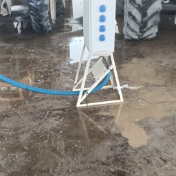 VoltMax - Biała, przenośna rozdzielnica budowlana z trzema niebieskimi gniazdami, podłączona niebieskim peszlem, stoi na metalowym stojaku w błotnistej hali z traktorami w tle.