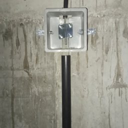 VoltMax - Szara puszka instalacyjna z metalowym uchwytem montażowym na surowej, betonowej ścianie z widocznymi zaciekami, obok czarny peszel mocowany metalowymi obejmami.