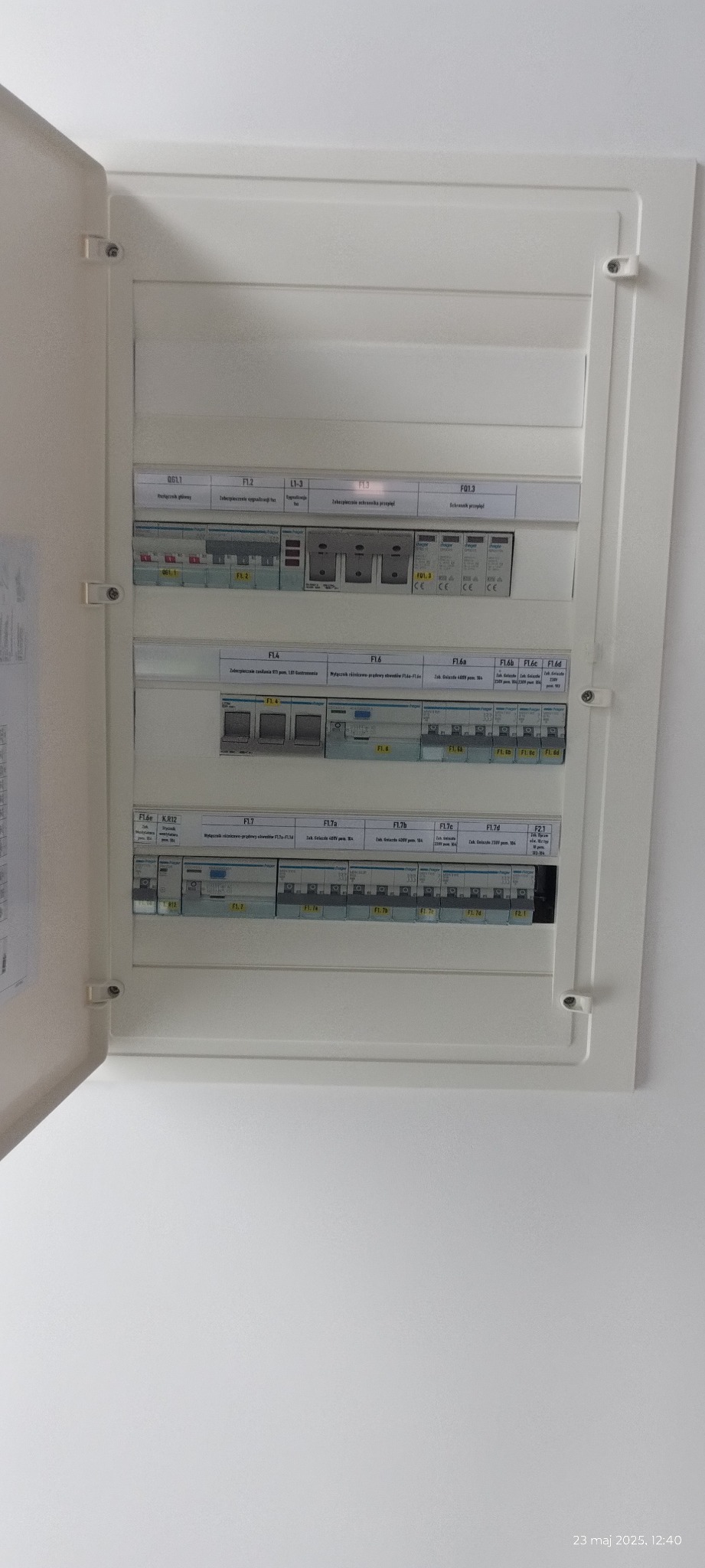 Otwarta skrzynka bezpiecznikowa z opisami i przełącznikami, widoczna na białej ścianie. Widok z bliska na elementy instalacji elektrycznej wewnątrz.