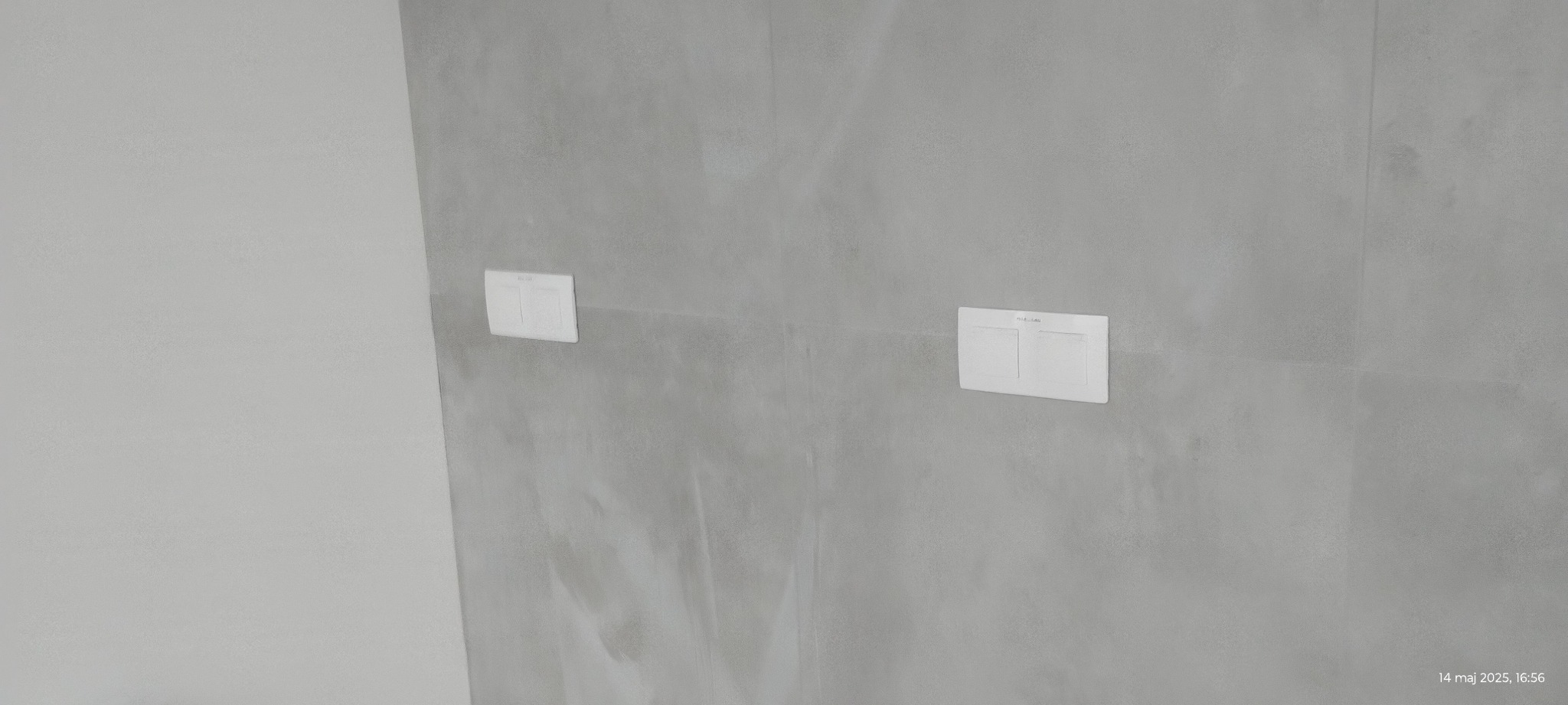 Dwa białe przyciski spłuczki wbudowane w szarą ścianę o teksturze betonu, widoczne z perspektywy lekko z dołu, z datą w prawym dolnym rogu.